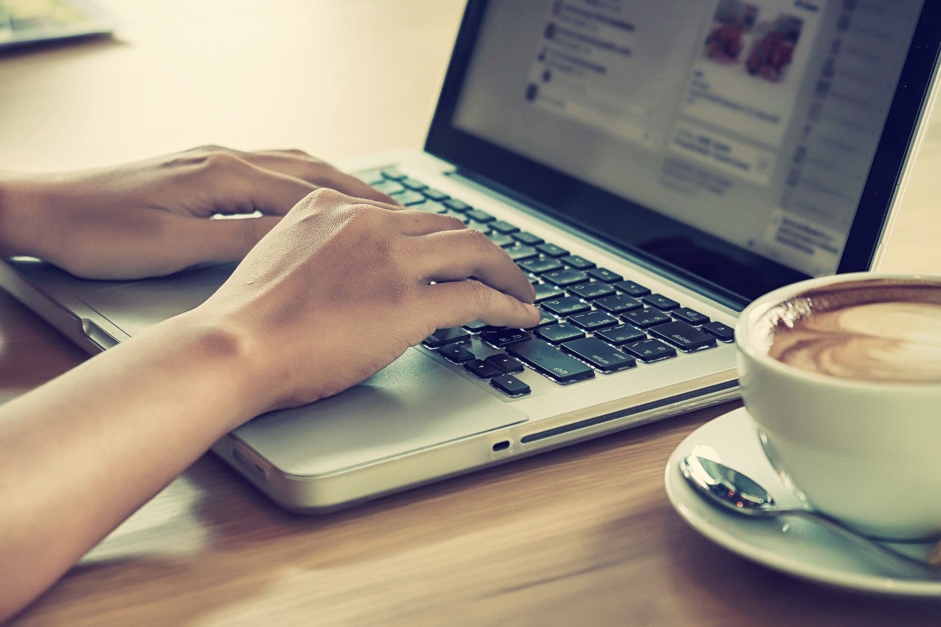 Person typing on laptop keyboard at desk with coffee cup and pen nearby