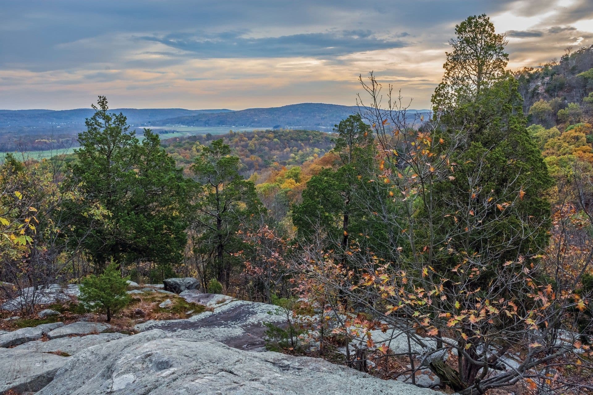 Scenic overlook of a forested valley with a lake in the distance, autumn foliage, rocky outcrop in foreground, and mountains under a cloudy sky