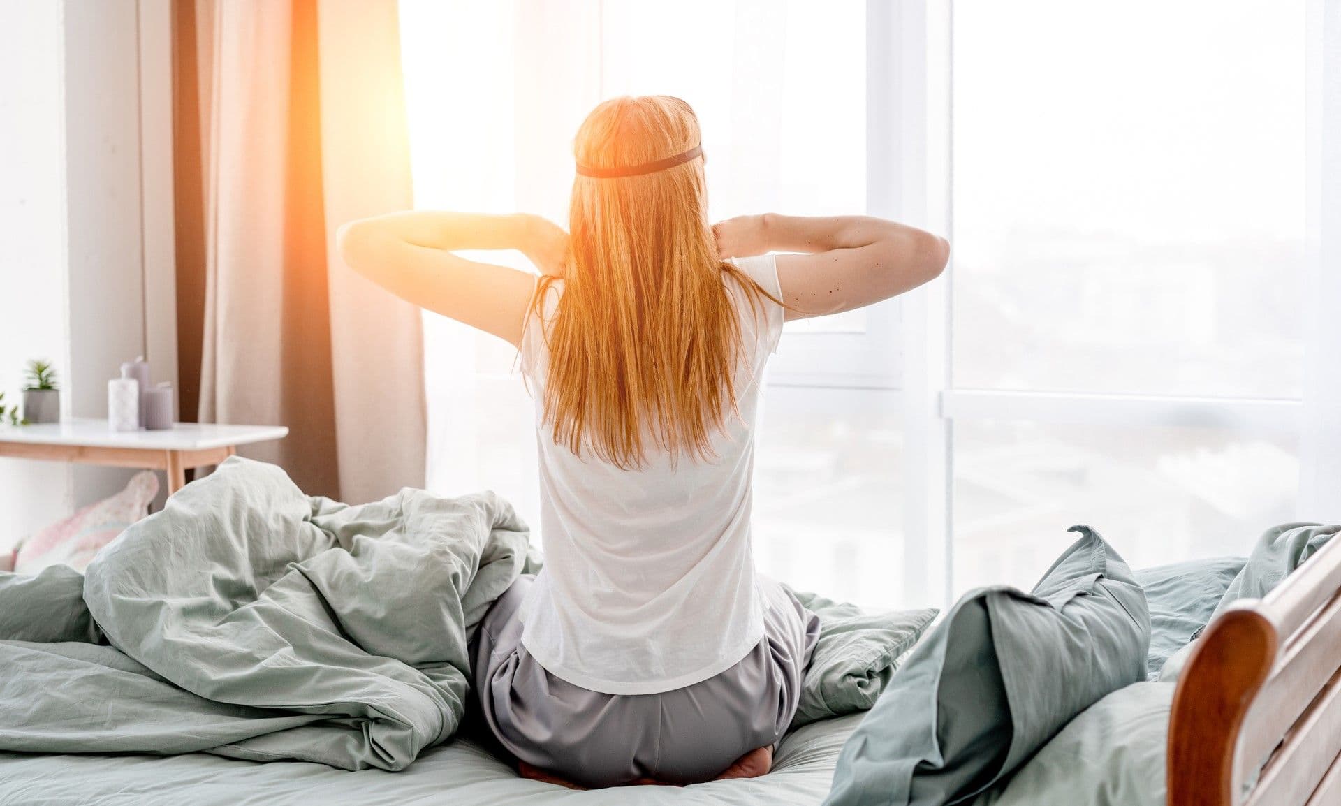 Woman with long red hair sitting on bed facing window with arms stretched behind head in relaxed morning pose