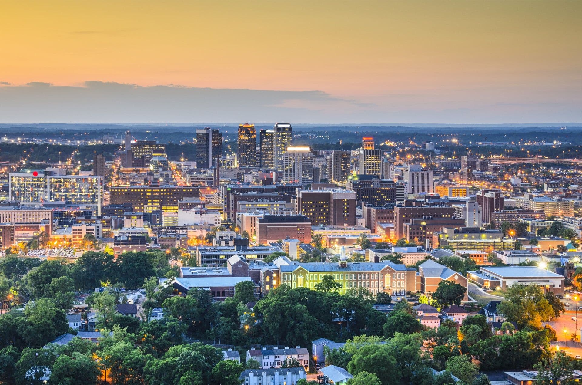 City skyline at dusk with illuminated buildings, green residential neighborhoods in foreground, and mountains visible on distant horizon