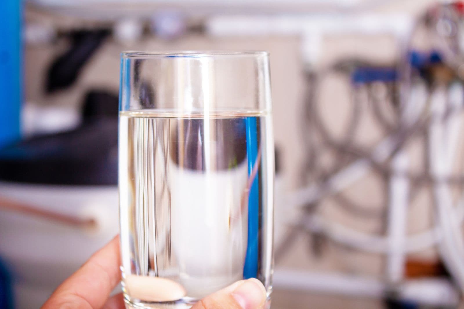 A hand holds a clear glass of water in a kitchen setting with blurred plumbing and filtration equipment in the background, conveying purity and freshness.