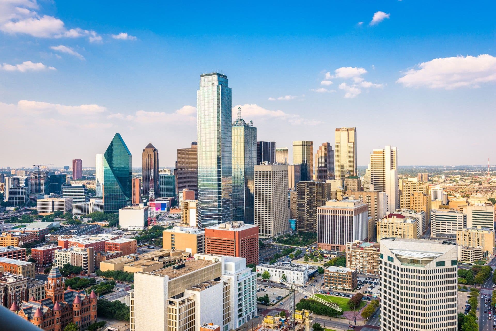 Dallas city skyline with distinctive tall buildings including the green geometric tower, surrounded by shorter historic and modern structures under blue sky with white clouds