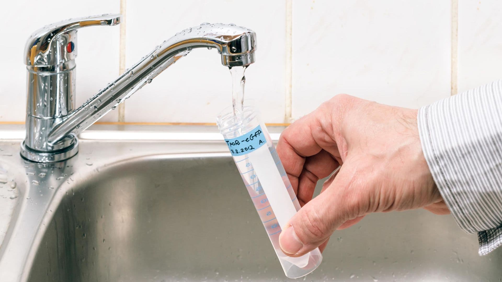 A hand holds a test tube under a running kitchen tap, collecting water for testing. The setting conveys a scientific and hygienic context.