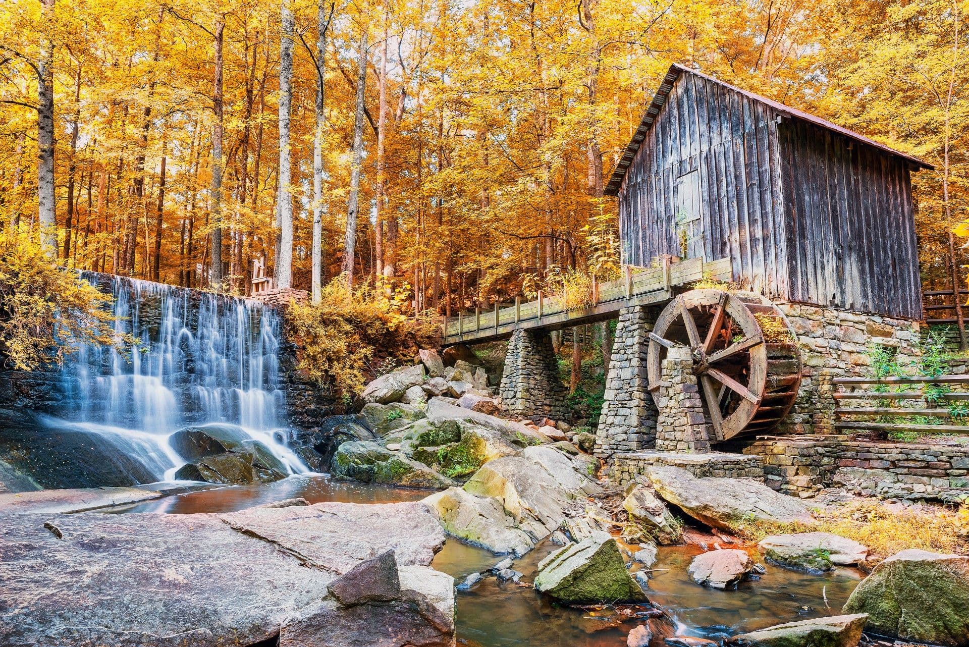 Historic grist mill with wooden water wheel beside cascading waterfall in autumn forest with golden foliage