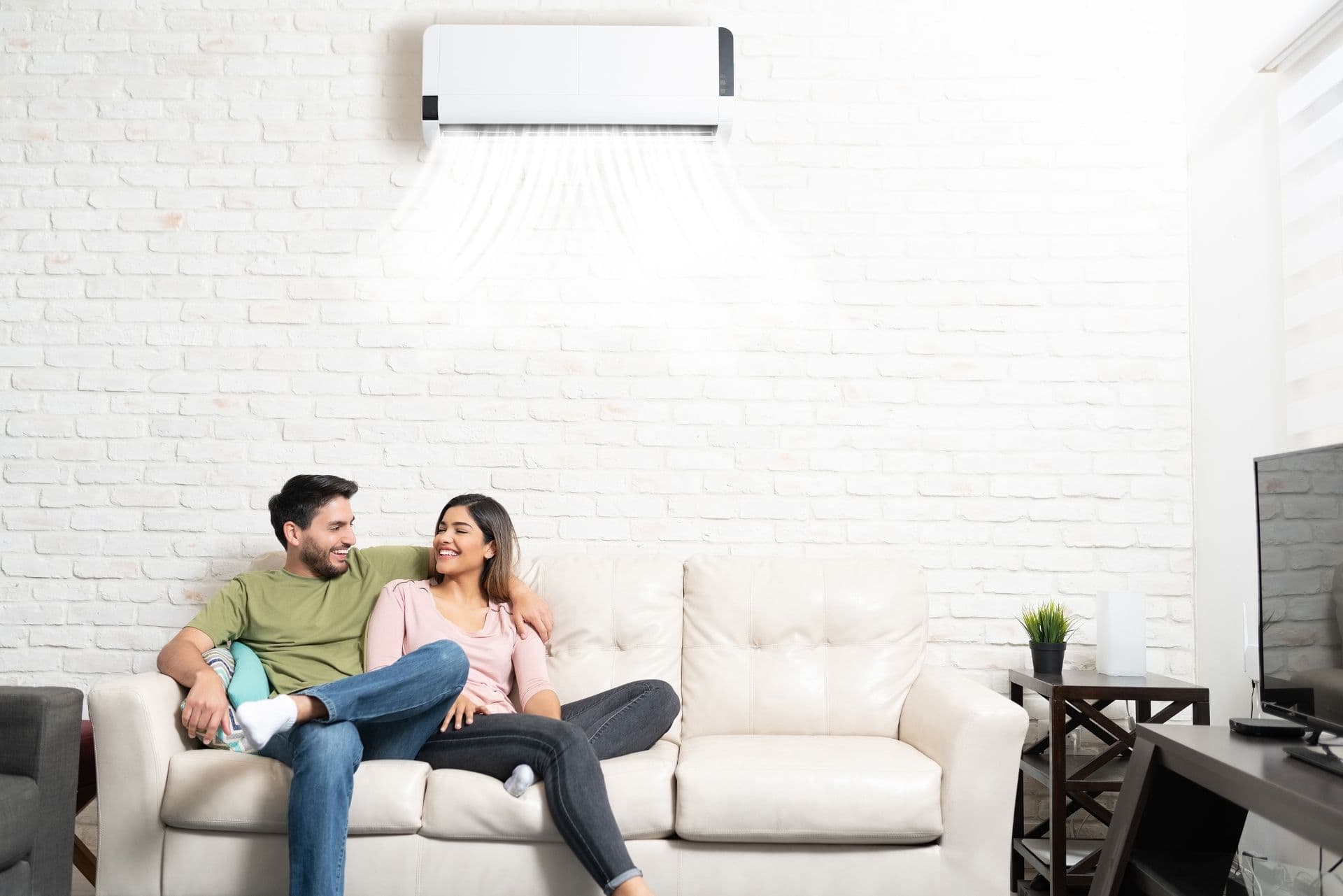 A smiling couple relaxes on a white sofa in a cozy living room with a white brick wall. An air conditioner above them emits cool air, creating a serene atmosphere.