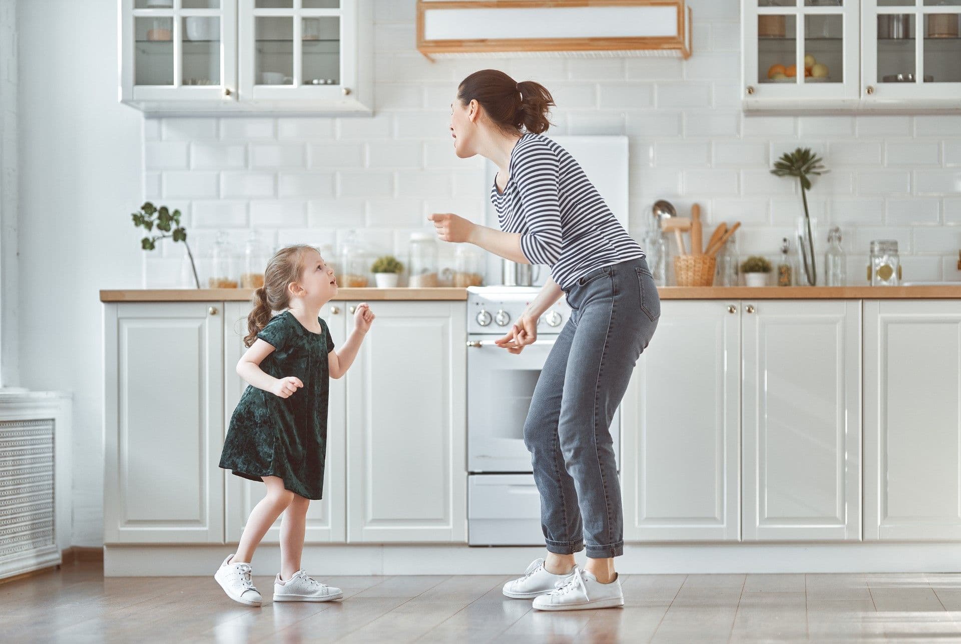 Woman and young child dancing together in a bright, modern kitchen with white cabinets and wooden countertops
