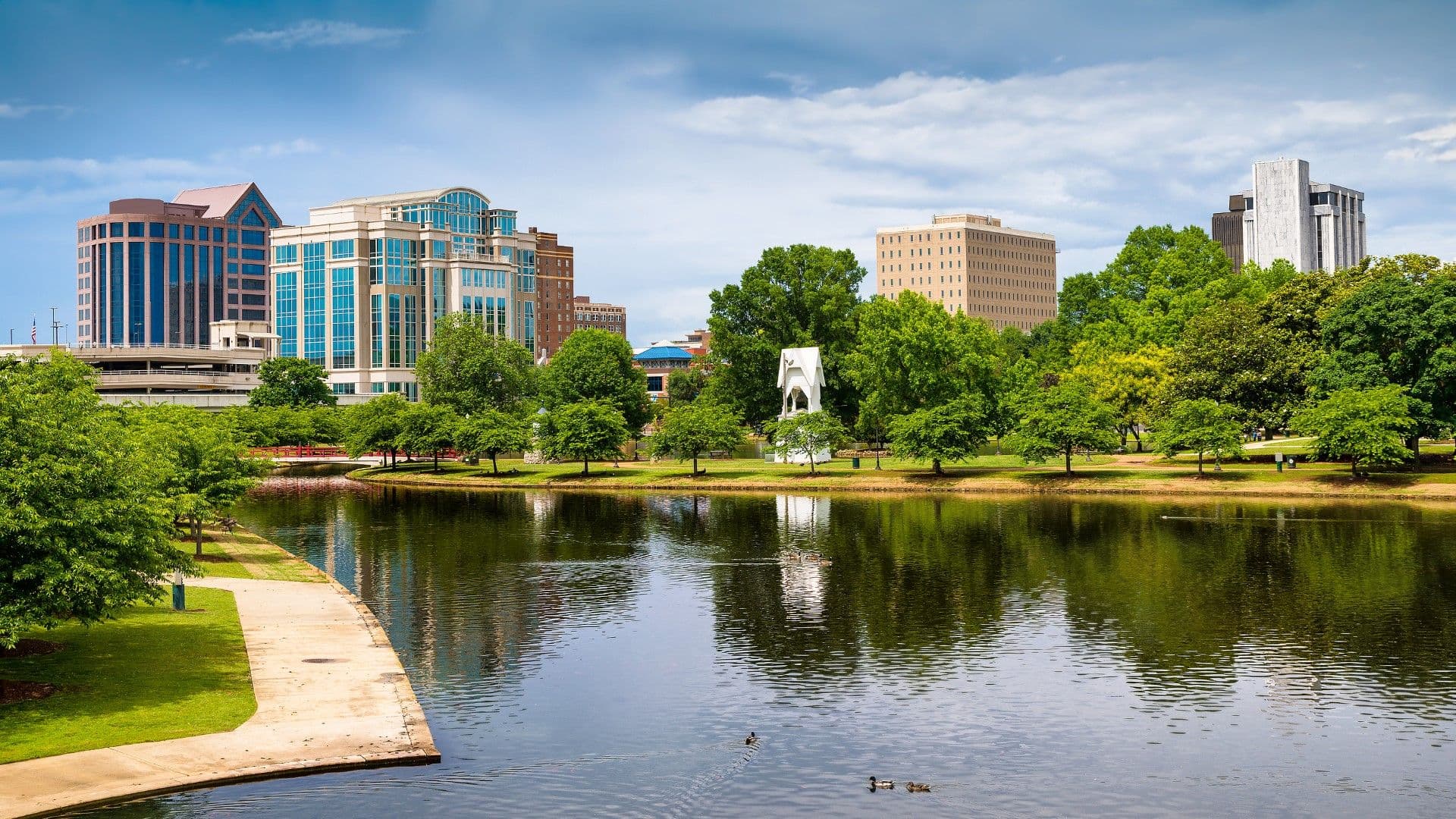 Urban riverfront park with ducks swimming in river, grassy lawn, mature trees, and downtown skyline featuring modern and historic buildings under blue sky
