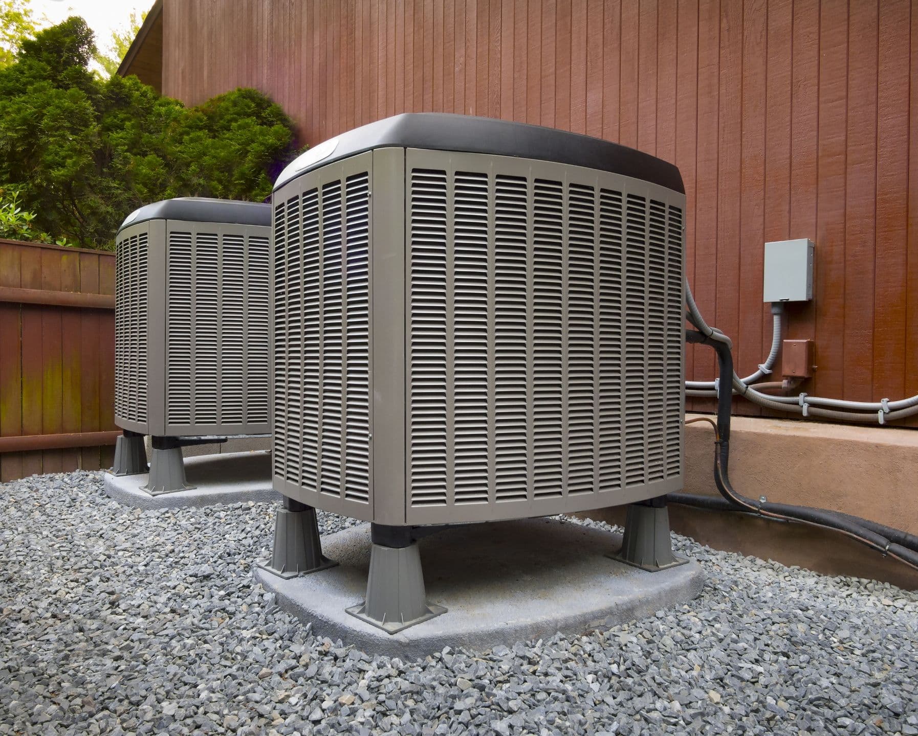 Two outdoor air conditioning units sit on concrete pads beside a wooden building, surrounded by gravel and connected with electrical and refrigerant lines.