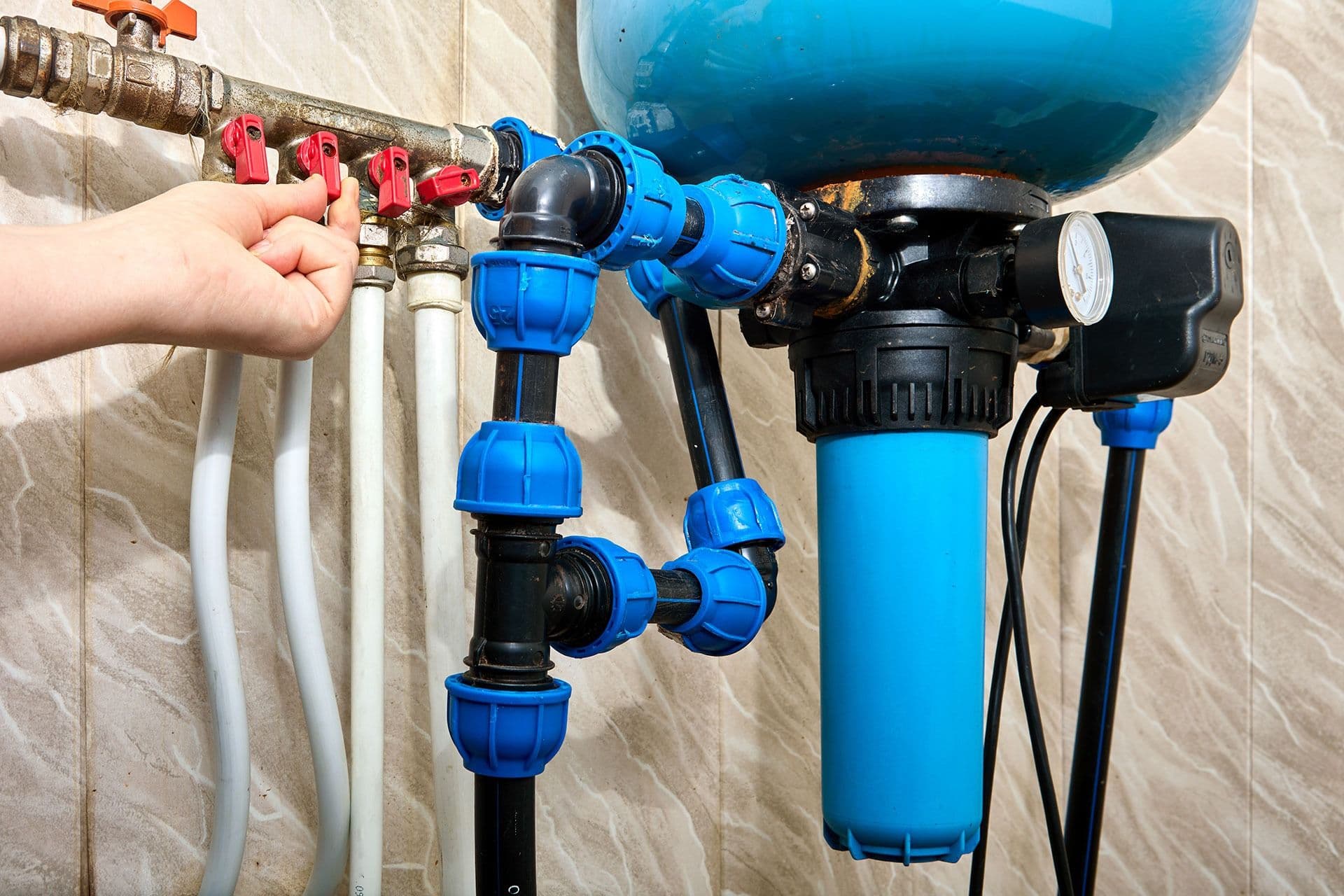 A hand adjusts a red valve on a complex water filtration system with blue pipes and a pressure gauge, set against a tiled wall.