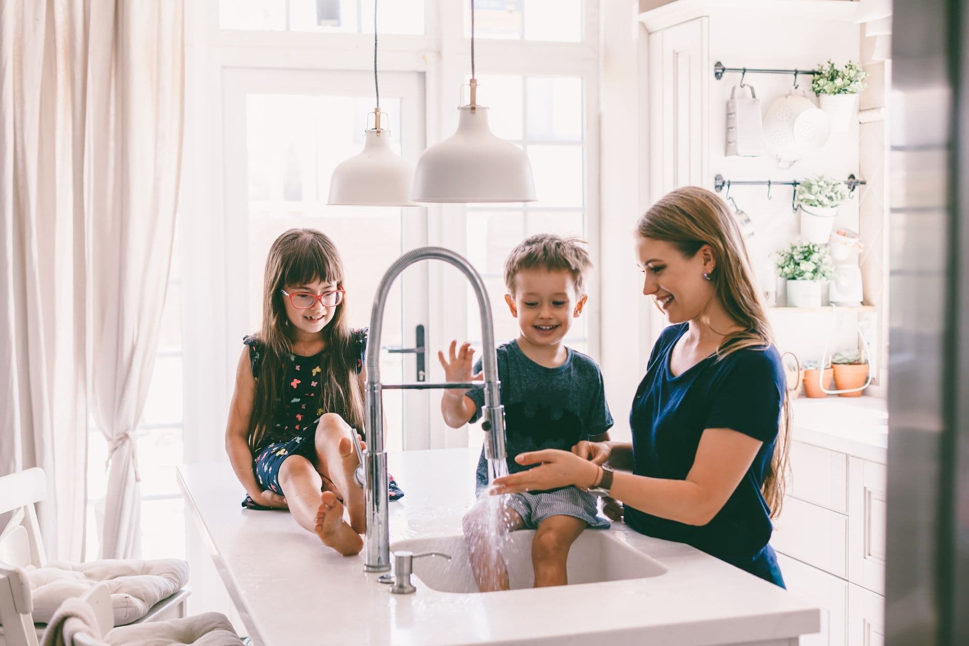 A woman and two children are playing with water at a kitchen sink. The children are sitting on the counter, smiling and splashing water, while the woman engages with them.