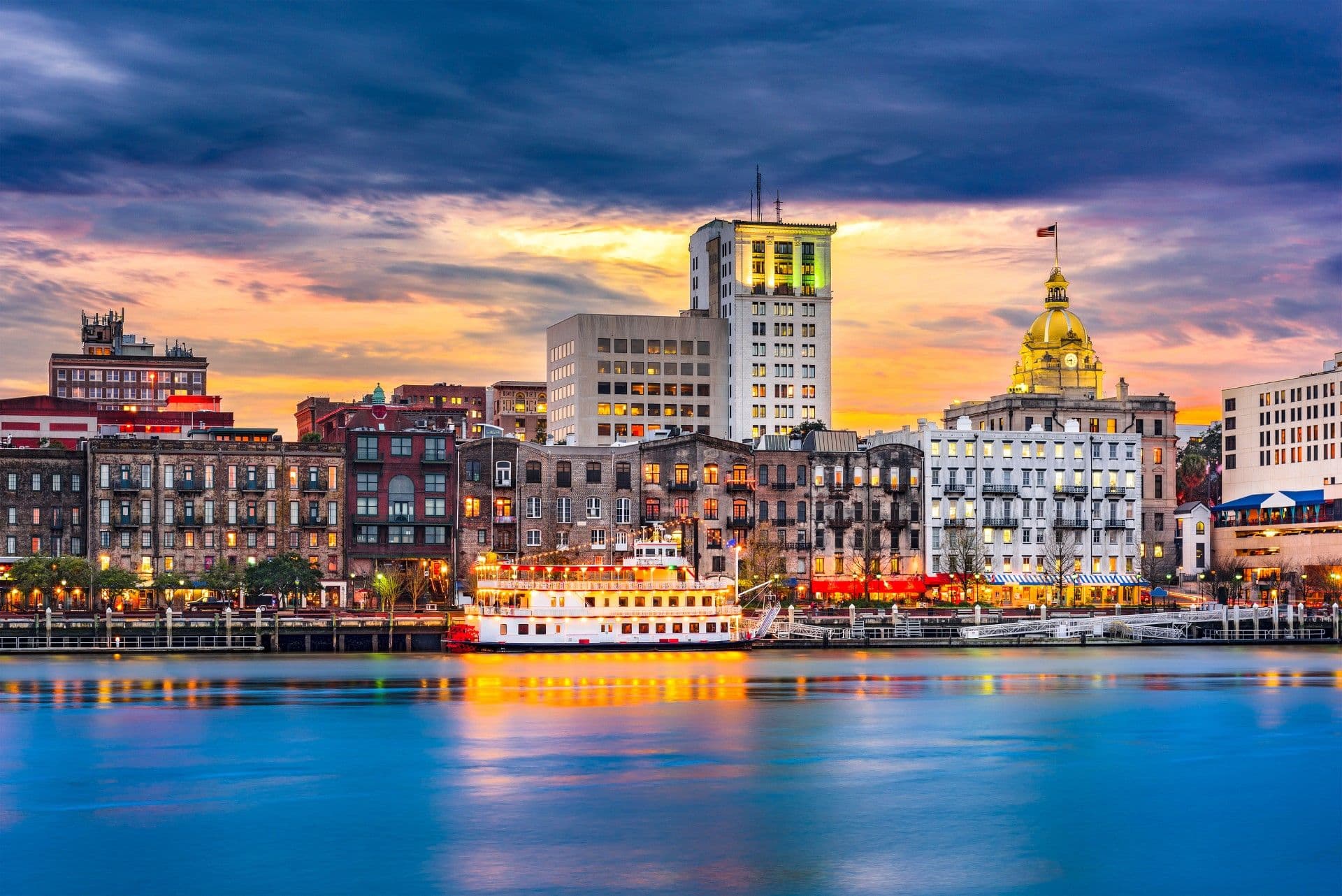 Savannah riverfront skyline at dusk with historic brick buildings, a domed courthouse with gold roof, and a red and white riverboat on the water