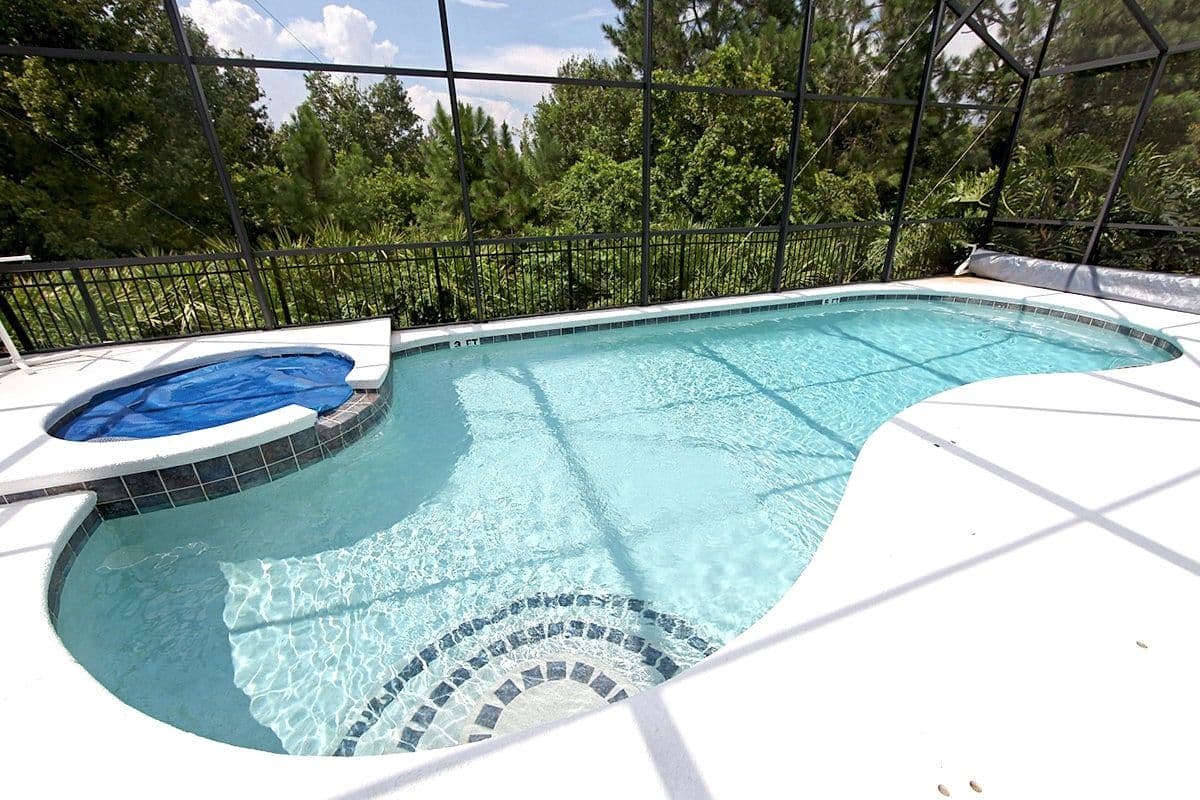 Enclosed backyard pool with sparkling blue water and curved design, bordered by lush green trees. A covered spa is adjacent, creating a serene atmosphere.