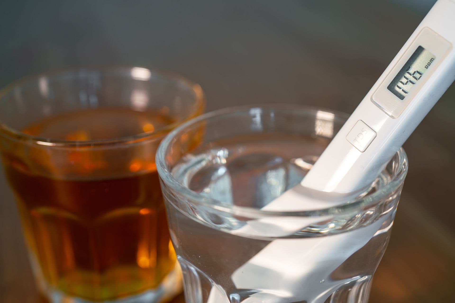 A digital water tester in a glass of clear water, with a second glass containing amber-colored liquid in the background on a wooden table.