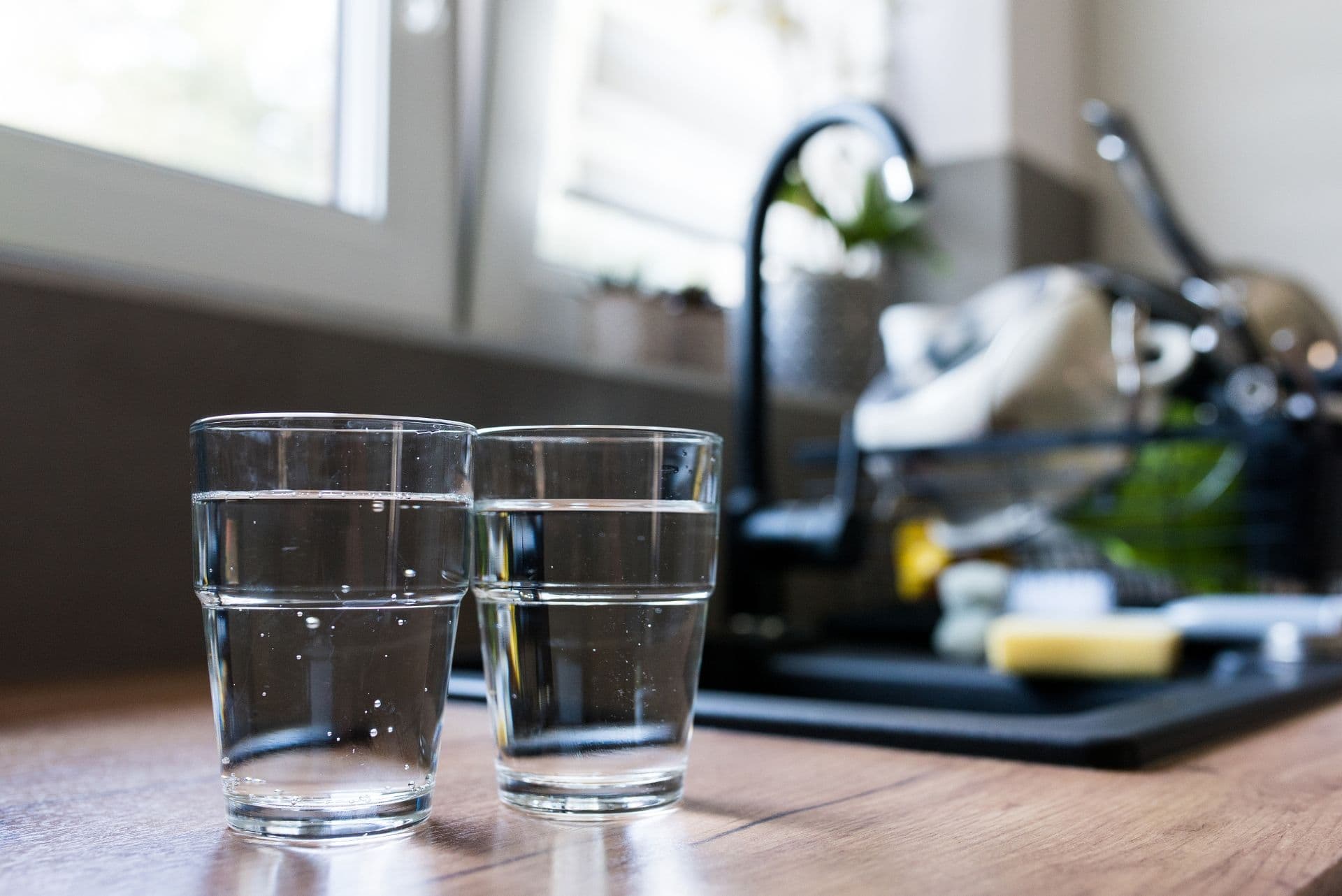 Two glasses of water sit on a wooden kitchen counter, with sunlight streaming through a nearby window. A dish rack is slightly blurred in the background.