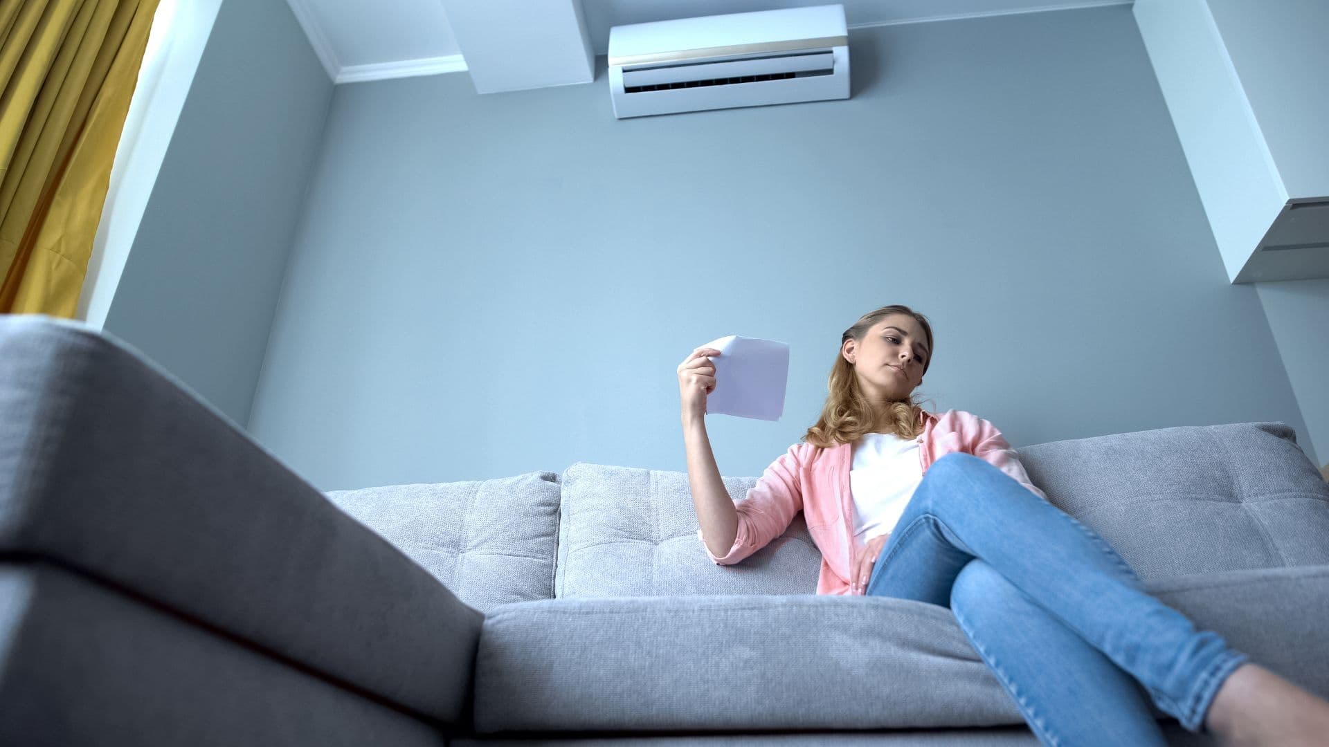 Woman sitting on a gray couch uses a paper to fan herself while looking up at a wall-mounted air conditioner in a modern living room.