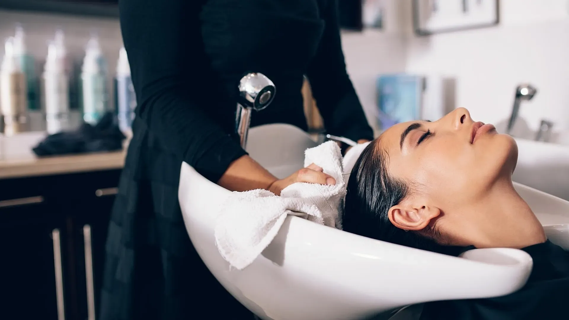 A woman relaxes at a salon while a stylist, dressed in black, gently washes her hair over a white sink. Various hair products are blurred in the background.