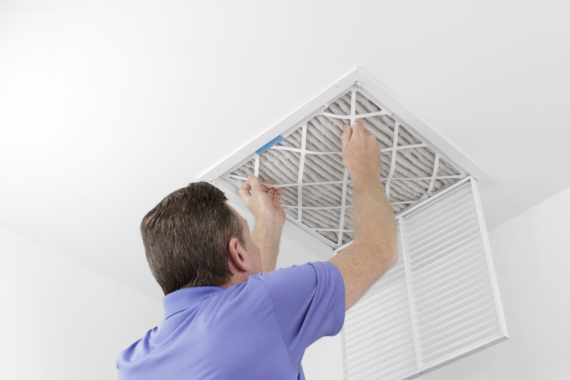 A person wearing a purple shirt reaches up to install or replace a pleated air filter into a white ceiling vent frame in a bright white room.