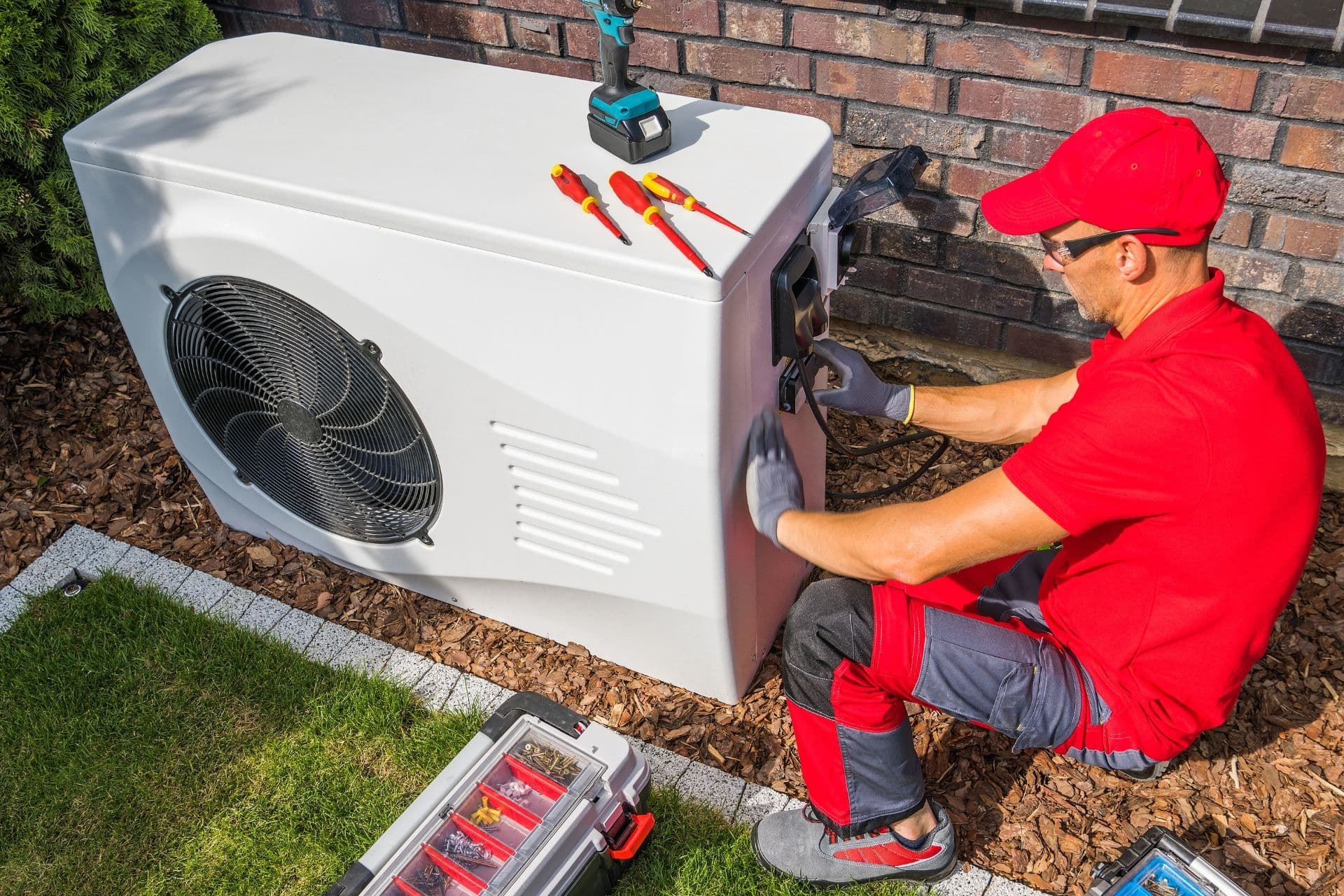 A technician in red uniform and cap fixes a white heat pump unit outdoors. Tools and a toolbox are nearby. The setting is a tidy lawn beside a brick wall.