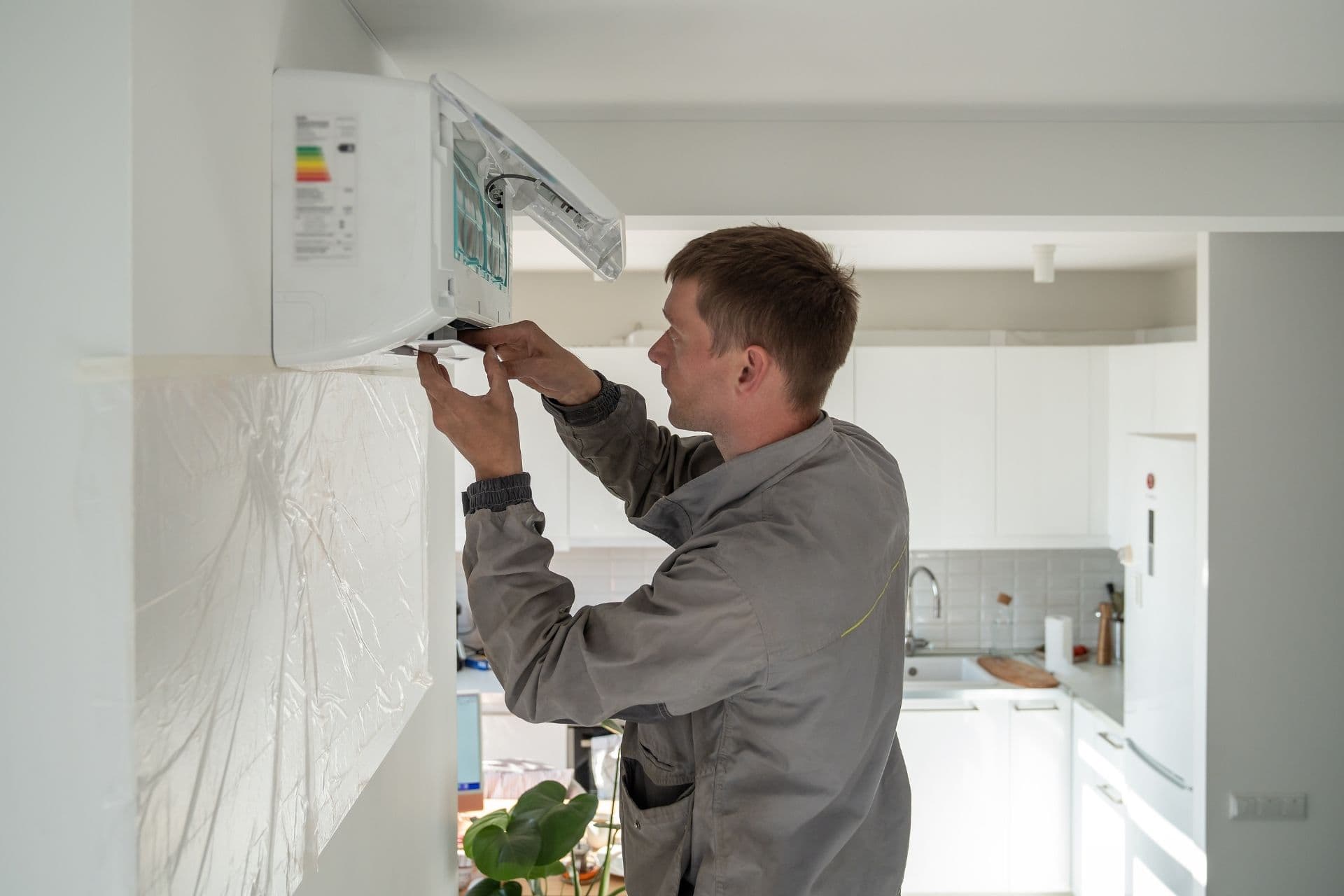 A technician in a grey uniform installs a white wall-mounted air conditioner in a modern kitchen, illustrating a focused, professional atmosphere.