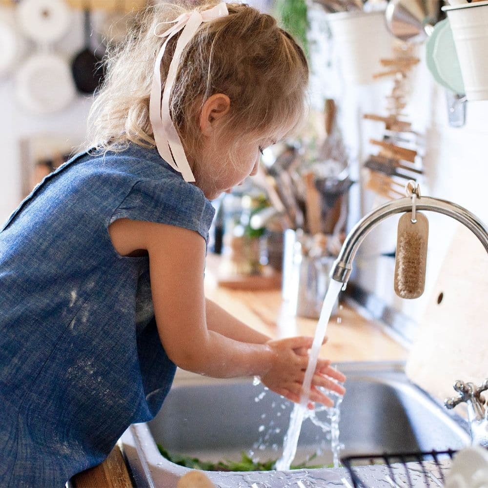 A cheerful little girl stands at a kitchen sink, scrubbing her hands with soap and water.