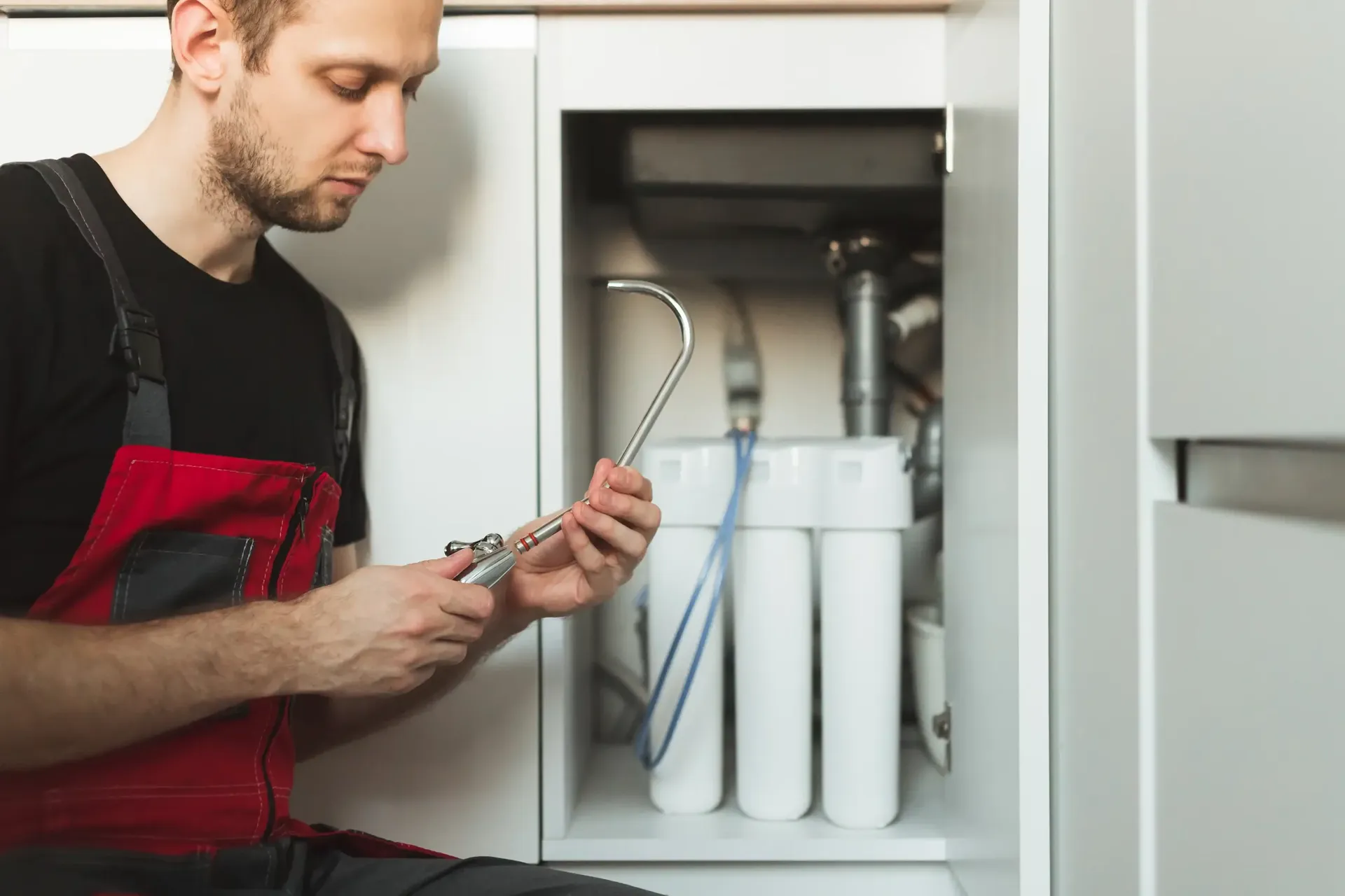 A plumber in a red apron examines a pipe wrench beside a cabinet with water filters. The scene conveys focus and professionalism in maintenance work.