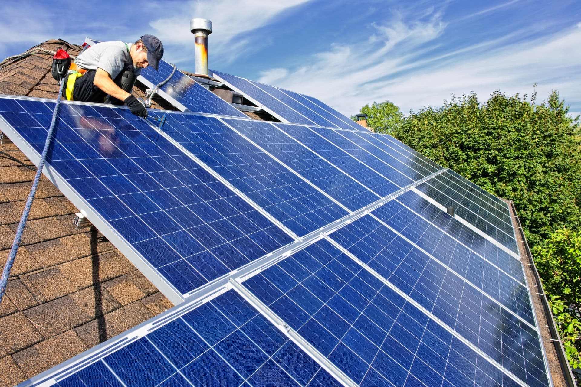 A worker installs solar panels on a sloped house roof, under a clear blue sky. Lush greenery surrounds the scene, conveying sustainability and eco-friendliness.